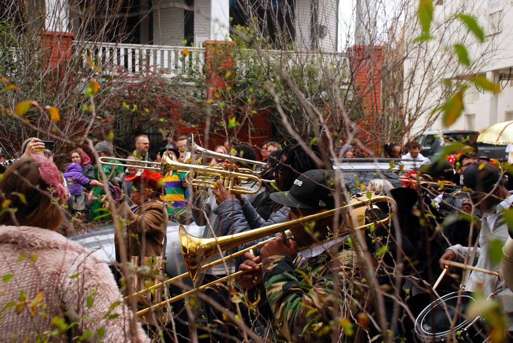 A brass band playing music during a parade in New Orleans street. There is a tambour, trumpet, and two trombone players.