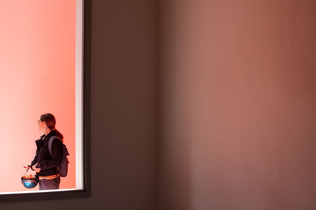 Through an empty windows frame inside an empty classroom, a women is looking outside. The ambient light is pink.