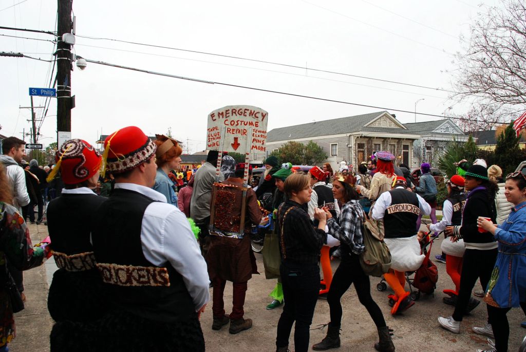 The redbean parade in New Orleans, with a lot of people walking in the street. One man stand in the middle with an improvise Emergency Costume repair portable workshop.