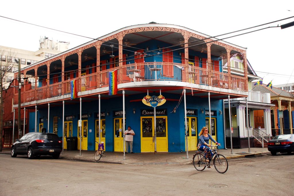 A blue restaurant selling hotdog called Dat Dog. There is pride flag hanging on the balcony. On the forefront a women on bike crossing the street.